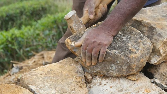 Male hands of indian stone worker cuts block of granite with a hammer for construction flowerbed. Adult unrecognizable stonemason hits a stone with sledgehammer in botanical garden. Slow motion