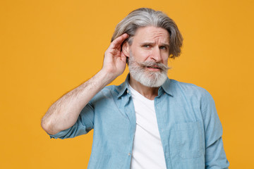 Curious elderly gray-haired mustache bearded man in casual blue shirt posing isolated on yellow...
