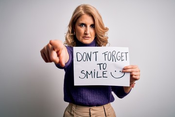 Middle age beautiful blonde woman holding banner funny positive message pointing with finger to the camera and to you, hand sign, positive and confident gesture from the front