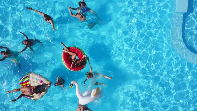 Aerial of friends having party in swimming pool with inflatable flamingo, swan, mattress. Happy young people wave hands at drone camera on luxury resort. View from above. Girls in bikini sunbathing.