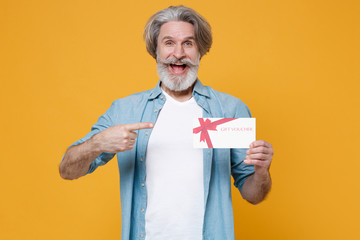 Excited elderly gray-haired mustache bearded man in casual blue shirt posing isolated on yellow background studio. People lifestyle concept. Mock up copy space. Point index finger on gift certificate.