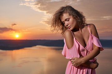 Girl with curly hair in pink dress on background of sunset.