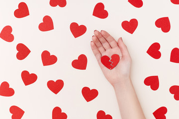 Pattern of red paper hearts surrounding woman's hand holding two parts of broken heart on the biege background
