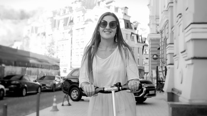 Obraz na płótnie Canvas Black and white portrait of beautiful smiling woman in sunglasses riding on electric scooter on city street