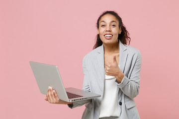 Smiling african american business woman in grey suit, white shirt isolated on pink background. Achievement career wealth business concept. Mock up copy space. Hold laptop pc computer showing thumb up.