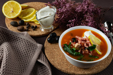 Soup Solyanka is a combined meat soup with smoked meat , various types of meat and tomato paste, serving dishes with sour cream, lemon and fresh herbs. Black olives on the table and a sprig of fresh f