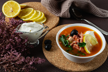 Soup Solyanka is a combined meat soup with smoked meat , various types of meat and tomato paste, serving dishes with sour cream, lemon and fresh herbs. Black olives on the table and a sprig of fresh f