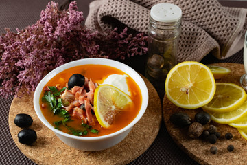 Soup Solyanka is a combined meat soup with smoked meat , various types of meat and tomato paste, serving dishes with sour cream, lemon and fresh herbs. Black olives on the table and a sprig of fresh f