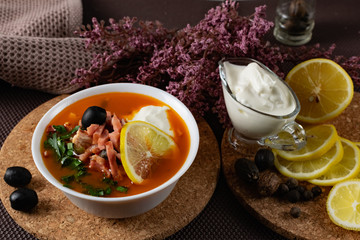 Soup Solyanka is a combined meat soup with smoked meat , various types of meat and tomato paste, serving dishes with sour cream, lemon and fresh herbs. Black olives on the table and a sprig of fresh f
