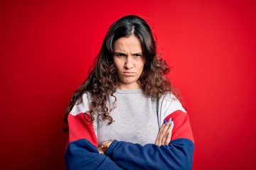 Young beautiful woman with curly hair wearing casual sweatshirt over isolated red background skeptic and nervous, disapproving expression on face with crossed arms. Negative person.