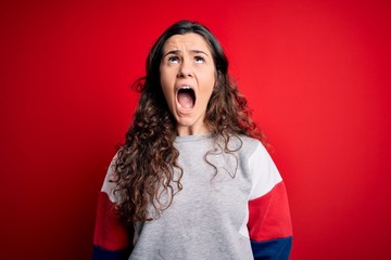 Young beautiful woman with curly hair wearing casual sweatshirt over isolated red background angry and mad screaming frustrated and furious, shouting with anger. Rage and aggressive concept.