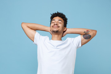 Smiling young african american guy in casual white t-shirt posing isolated on pastel blue wall background studio portrait. People lifestyle concept. Mock up copy space. Sleep with hands behind head.