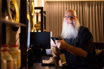 Happy mature bald bearded man using phone while working overtime at home late at night