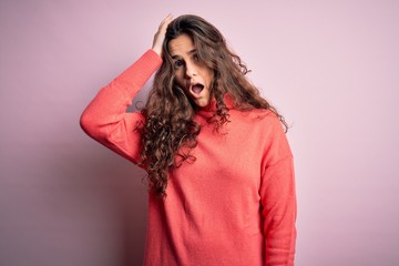 Young beautiful woman with curly hair wearing turtleneck sweater over pink background surprised...