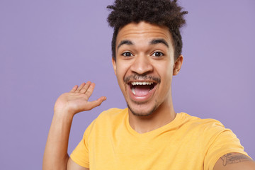 Close up of excited young african american guy in casual yellow t-shirt posing isolated on pastel violet background. People lifestyle concept. Doing selfie shot on mobile phone, pointing hand aside.