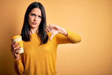 Young woman with blue eyes holding cup of coffee standing over yellow background with angry face,...