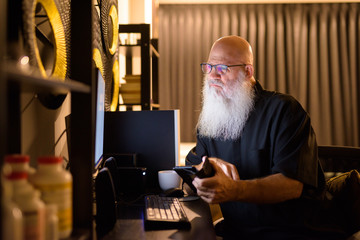 Mature bald bearded man thinking and using phone while working overtime at home late at night