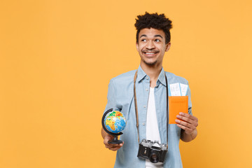 Smiling young african american guy in blue shirt with photo camera traveling abroad isolated on yellow background. Air flight journey concept. Mock up copy space. Hold passport ticket world globe.