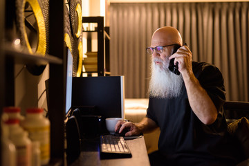 Mature bald bearded man talking on the phone while working overtime at home late at night