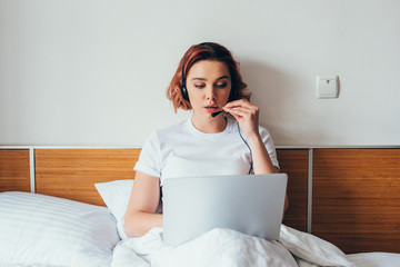 young woman making video call with headset and laptop in bed during quarantine