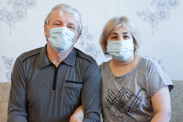 Protective masks are of faces of elderly people, senior couple sitting on a sofa at home