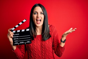 Young director woman with blue eyes making movie holding clapboard over red background very happy...