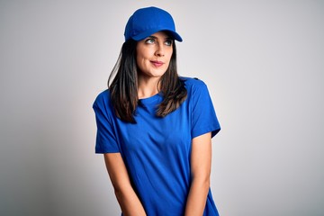 Young delivery woman with blue eyes wearing cap standing over blue background smiling looking to the side and staring away thinking.