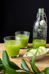 Top view of two glasses with cucumber juice and bottle, bowl with slices, mint and unfocused green branch, on wooden table and black background, vertical