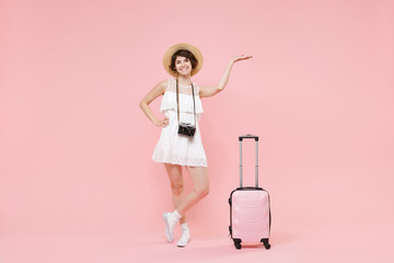 Smiling tourist girl in summer dress hat with photo camera suitcase isolated on pink background. Female traveling abroad to travel weekend getaway. Air flight journey concept. Pointing hand aside up.