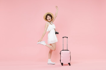 Funny tourist girl in summer dress hat with photo camera suitcase isolated on pink background. Female traveling abroad to travel weekend getaway. Air flight journey concept. Rising spreading hands.