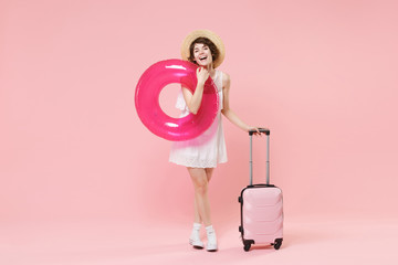 Cheerful young tourist woman in summer white dress hat with suitcase isolated on pink background. Female traveling abroad to travel weekends getaway. Air flight journey concept. Hold inflatable ring.