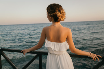 Fototapeta na wymiar Beautiful young smiling girl in the white dress in greek style walking along the sea promenade. Summer, sunset time. Bride wedding at sea 