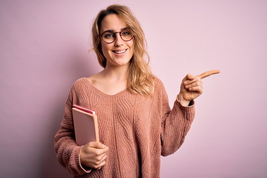 Young beautiful blonde woman wearing glasses holding notebook over pink background very happy pointing with hand and finger to the side