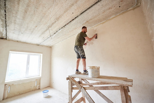 Builder man is standing on a wooden stand and working with a spatula with plaster on the wall against a window. Puttying the walls indoors. A guy with a beard in a t-shirt and jeans is smeared paint