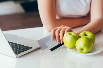 cropped view of woman shopping online with laptop and credit cards on kitchen with apples during self isolation
