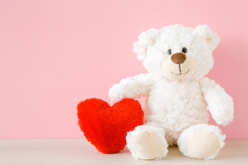 Smiling white teddy bear sitting and holding red soft heart at pastel pink wall. Front view....
