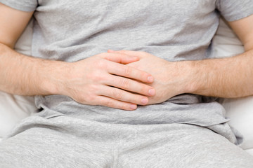 Young man sitting on bed. Hands touching belly. Suffering from abdominal pain at home. Stomach ache problems and solution concept. Closeup. Front view.