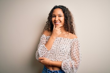 Young beautiful woman with curly hair wearing casual t-shirt standing over white background looking confident at the camera smiling with crossed arms and hand raised on chin. Thinking positive.