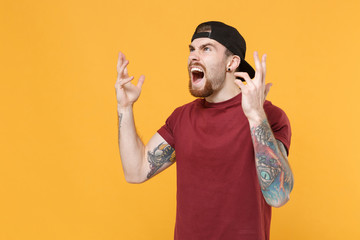 Shocked angry young bearded tattooed man guy in casual t-shirt, black cap isolated on yellow...