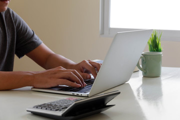 Naklejka premium An Asian man uses a laptop at home while sitting at a wooden table. Man hands typing on a notebook keyboard. The concept of young people working at mobile devices.