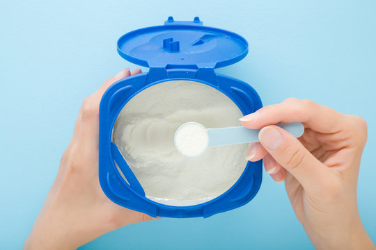 Young Mother Hand Taking White Powder Of Milk Formula With Spoon. Opened Container On Light Blue Table Background. Baby Feeding Concept. Closeup. Point Of View Shot. Top Down View.