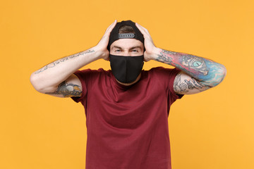 Displeased young tattooed man guy in casual t-shirt cap black face mask posing isolated on yellow background studio portrait. People emotions lifestyle concept. Mock up copy space. Put hands on head.