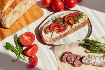 Bruschetta from fresh bread with cream cheese,  fouet, tomato slices and arugula in handmade plate on white wooden background. Italian or Spanish cuisine appetizer. Natural sun lighting