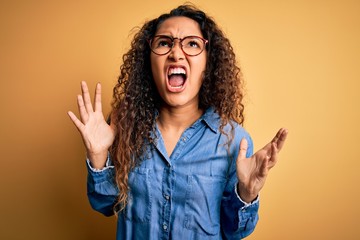 Beautiful woman with curly hair wearing casual denim shirt and glasses over yellow background crazy...