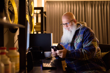 Mature bald bearded hipster man using phone while working overtime at home late at night