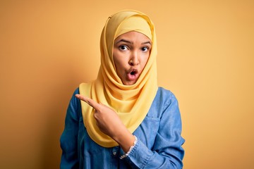 Young beautiful woman with curly hair wearing arab traditional hijab over yellow background Surprised pointing with finger to the side, open mouth amazed expression.