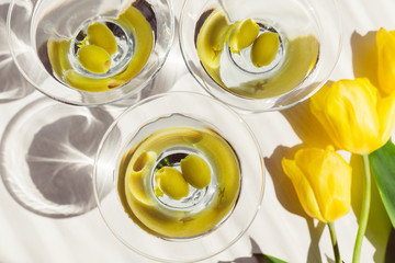 Top view of three glasses of martini cocktail with green olives for hen-party. White background with yellow tulips. Sun lighting