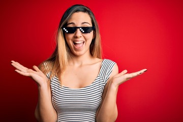 Young beautiful brunette woman wearing funny thug life sunglasses over red background celebrating crazy and amazed for success with arms raised and open eyes screaming excited. Winner concept