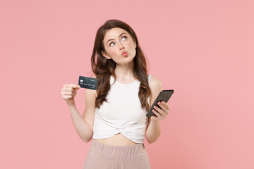 Pensive young brunette woman girl in light casual clothes posing isolated on pastel pink background in studio. People lifestyle concept. Mock up copy space. Using mobile phone, hold credit bank card.