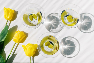 Fresh cold vodkatini or martini cocktail with green olives on white wooden table background with yellow tulips. Party time